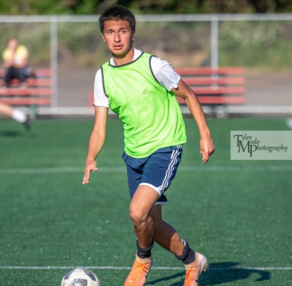 Soccer player in green vest dribbles ball on a turf field.