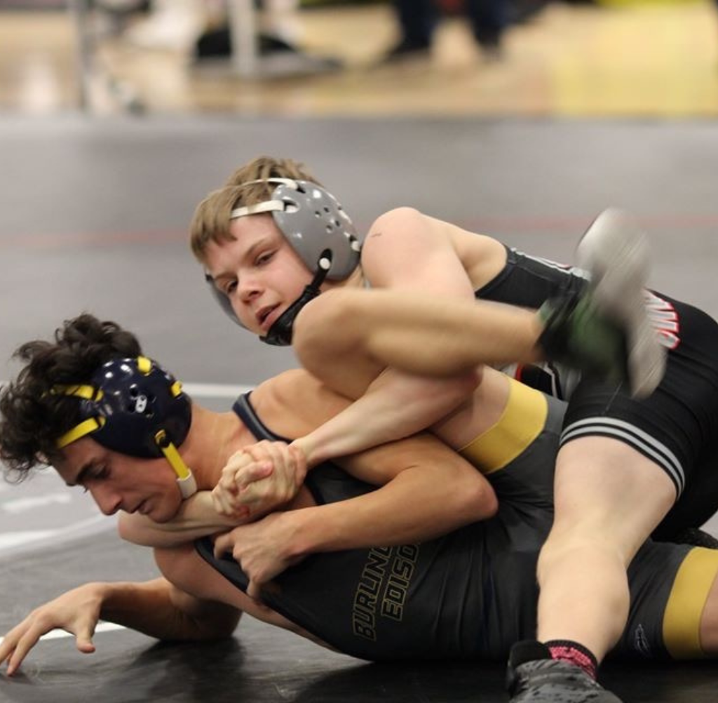 Two wrestlers in action, one on top, grappling on mat. Both wearing headgear and singlets.