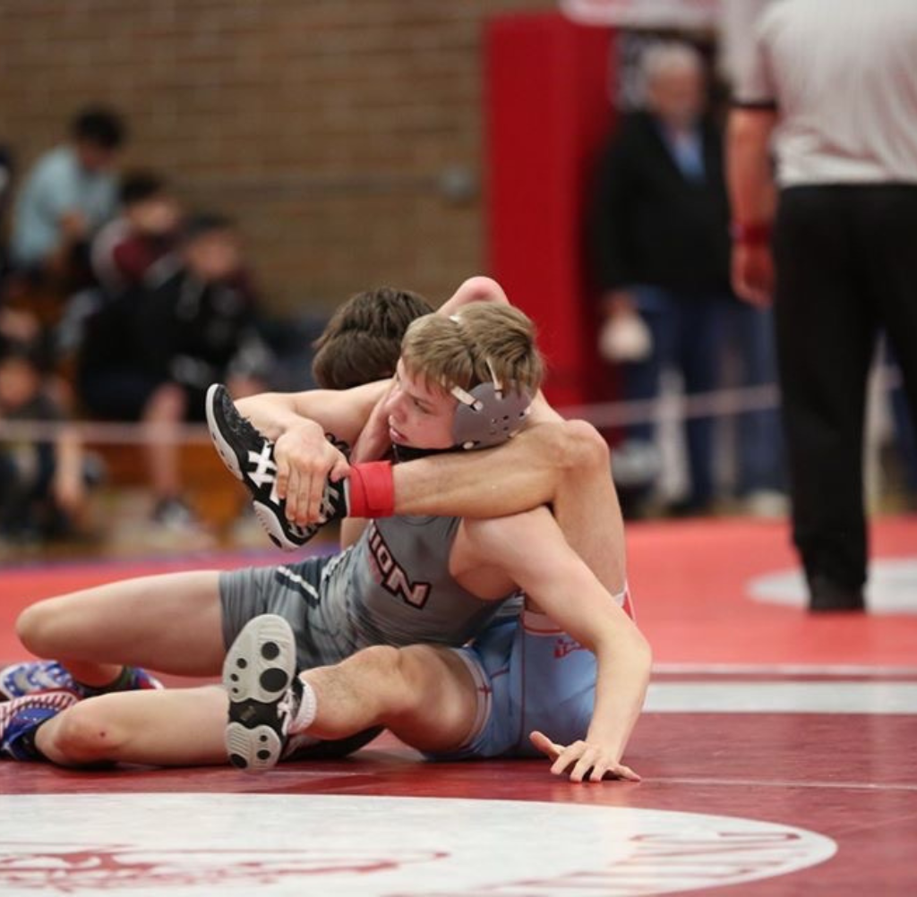 Two wrestlers in a match, one on top, red/gray singlet. Wrestling mat.