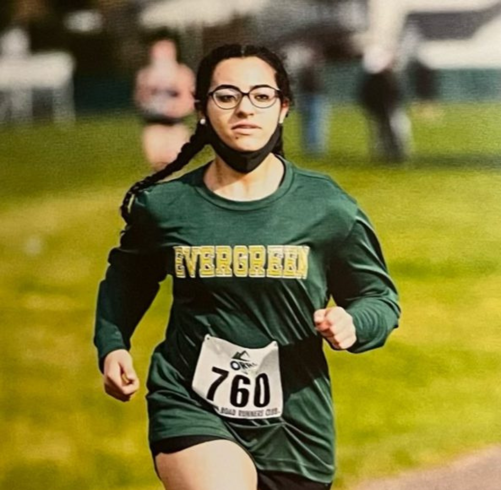 Young woman running a cross country race, wearing a green Evergreen shirt and race bib.