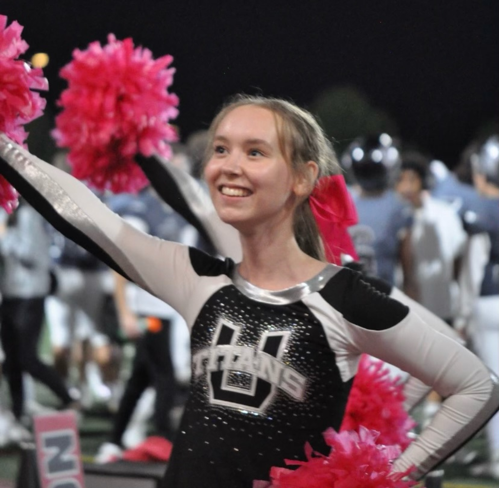 Cheerleader in black and white uniform with pink pom-poms, smiling at a football game.