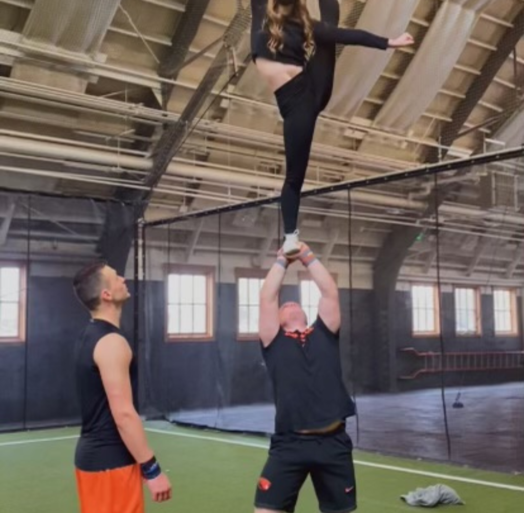 Two men lift a woman doing a split high in the air. She is in a black outfit. Indoors with a net.
