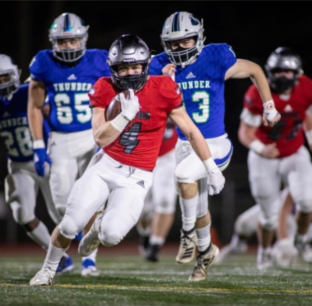 Football player in red jersey runs with ball, pursued by blue-clad players. Night game.