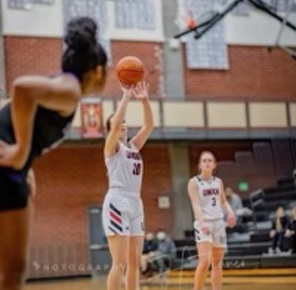 Basketball player in white jersey shooting the ball; two other players watch in a gymnasium.