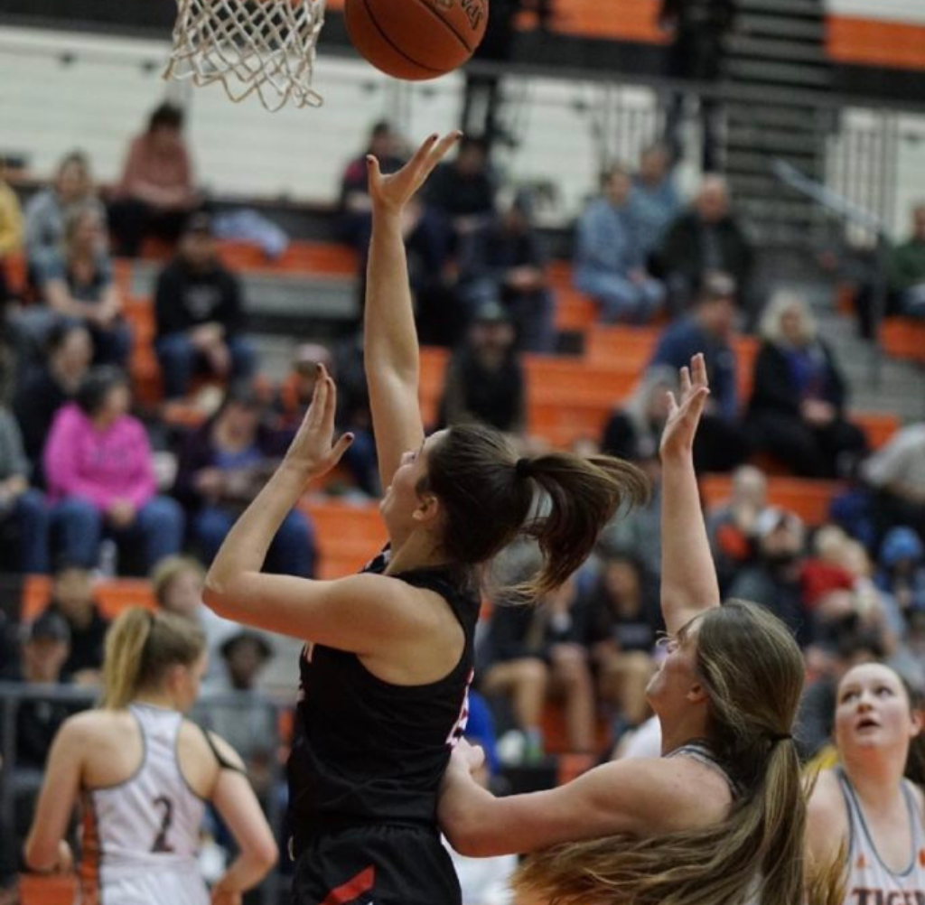 Basketball player shoots the ball during a game, challenged by a defender. Crowd in the background.