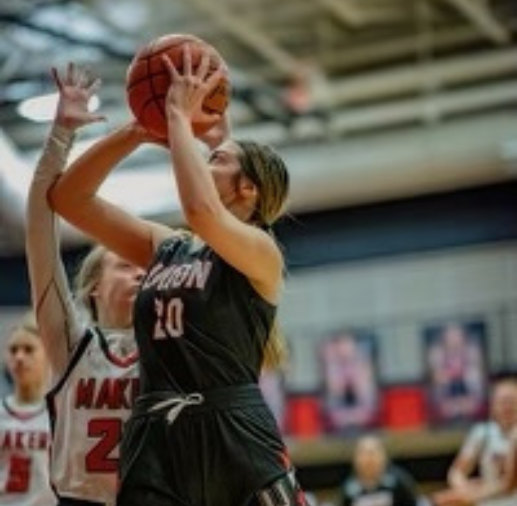 Basketball player in black jersey shoots the ball, closely guarded by a player in red jersey.