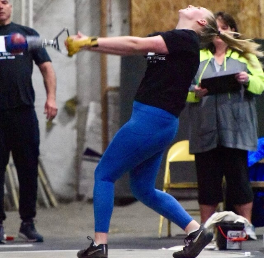 Woman in blue leggings throws a hammer, indoors. Leaning back, blonde hair flying.