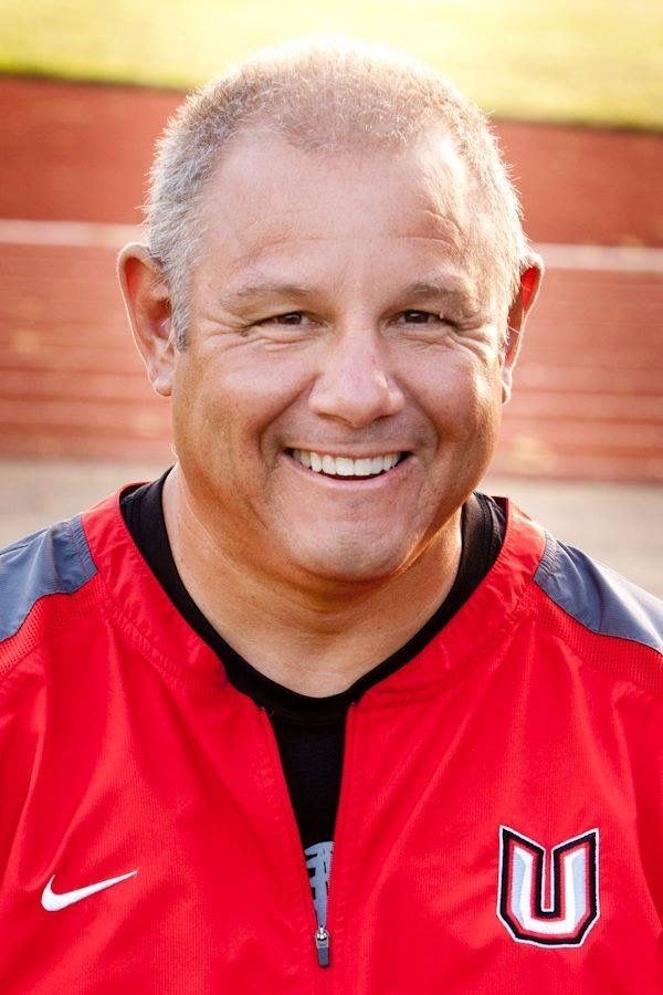 Smiling bald man in red jacket, posing outdoors near a track.