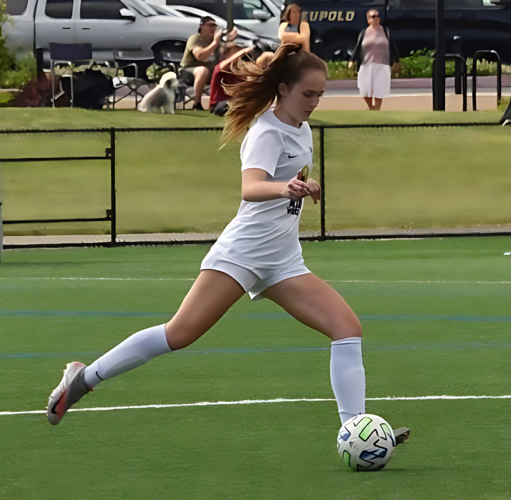 Female soccer player kicking the ball on a green field. White uniform, action shot, sunny day.