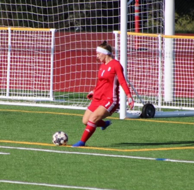 Rego Scholarship Soccer player in red uniform kicking ball on a green field. Goal net in the background.
