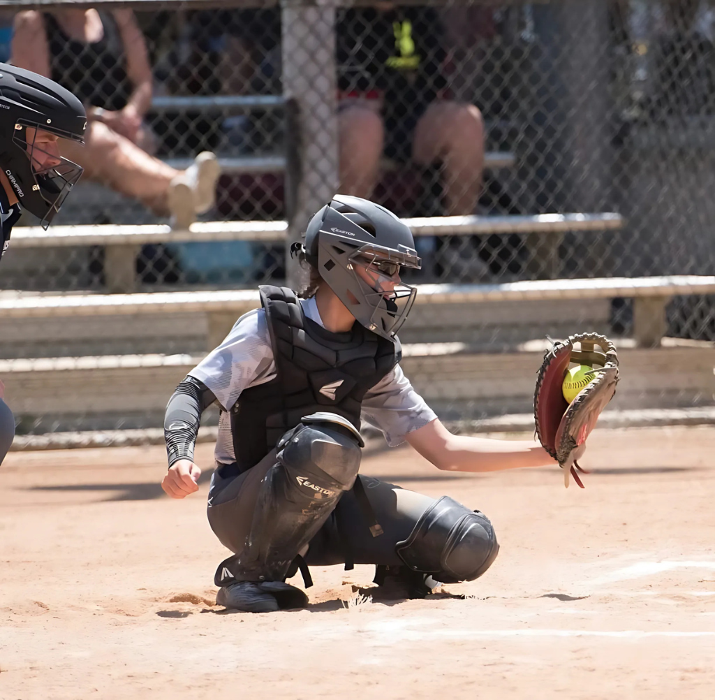 Softball catcher in gray and black uniform catches a yellow ball.