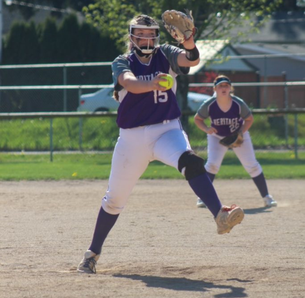 Softball pitcher in purple uniform, #15, winds up to throw. Another player in the background.