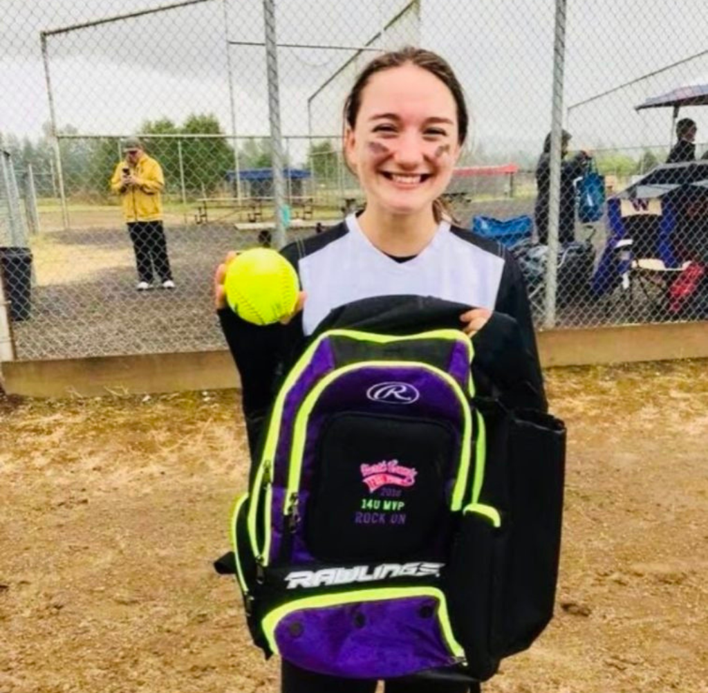 Softball player smiles, holding ball and purple Rawlings backpack; outdoors, overcast day.