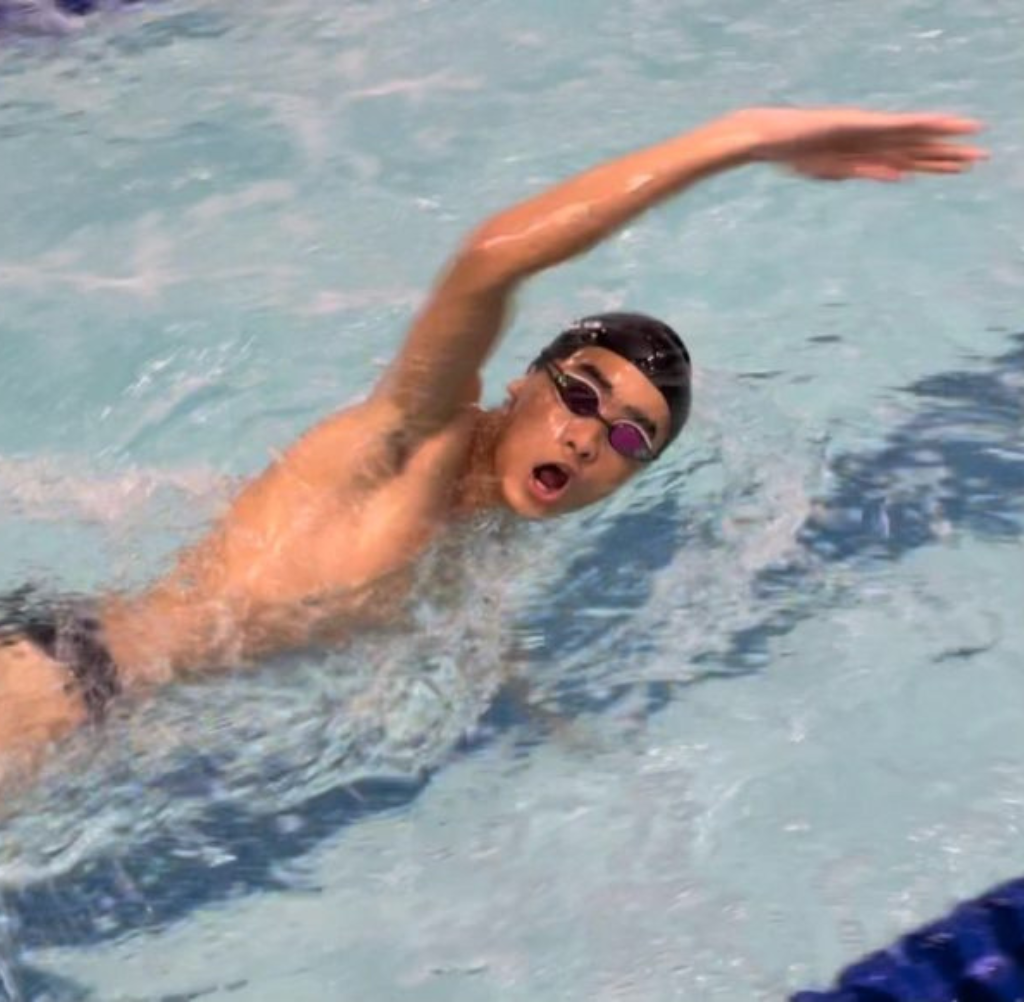Swimmer in a pool performing freestyle stroke, wearing goggles and a black cap, open mouth, arm extended.