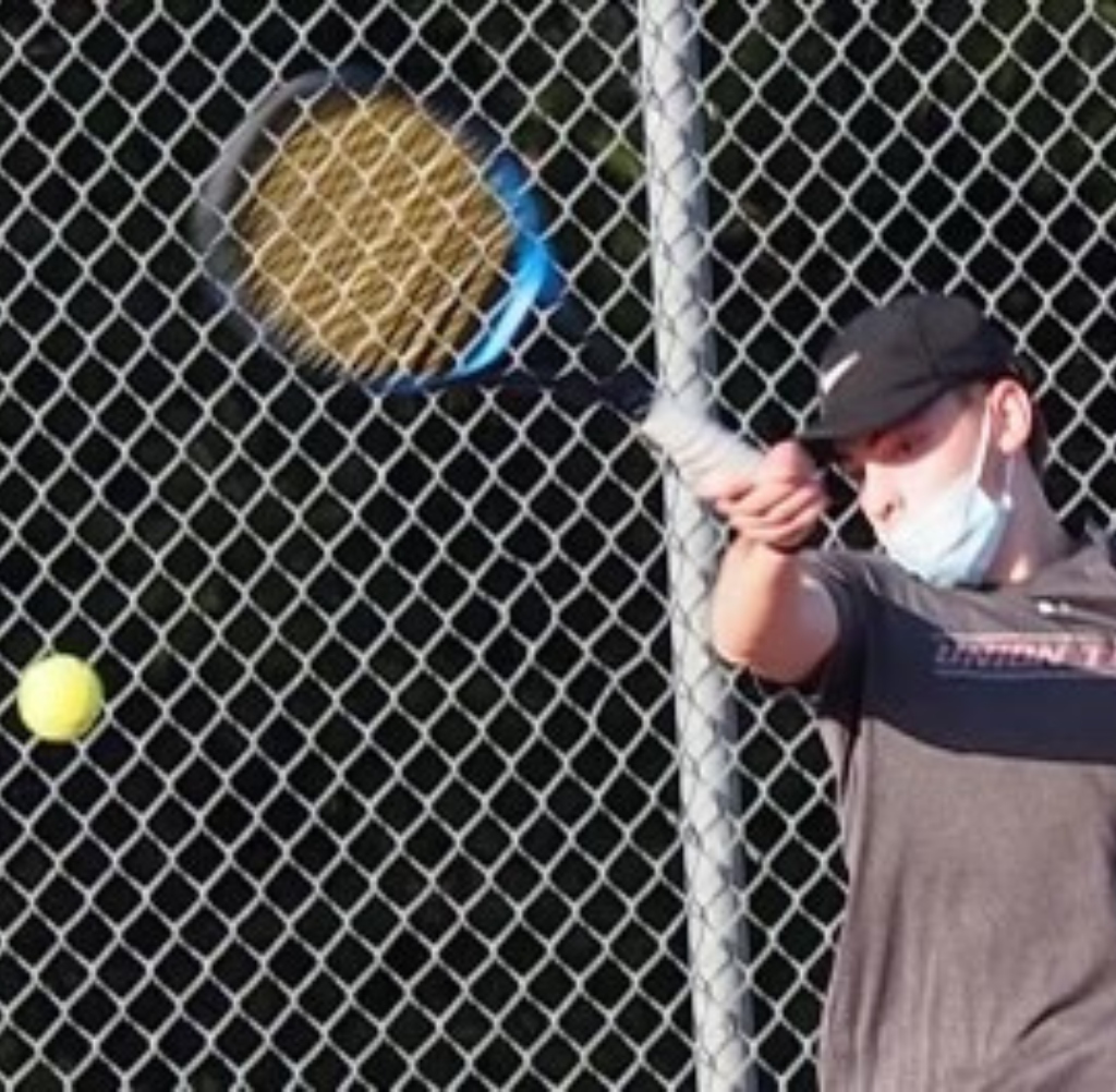 Man in a mask playing tennis; holding racket, ball in air, chain-link fence in background.