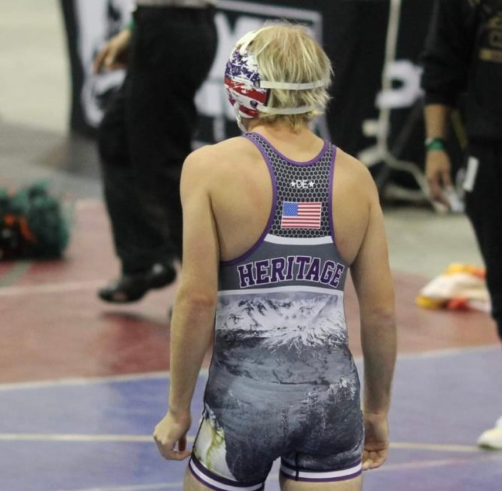 Wrestler in a Heritage singlet, with a mountain scene design, wearing a patriotic mask, stands on a mat.