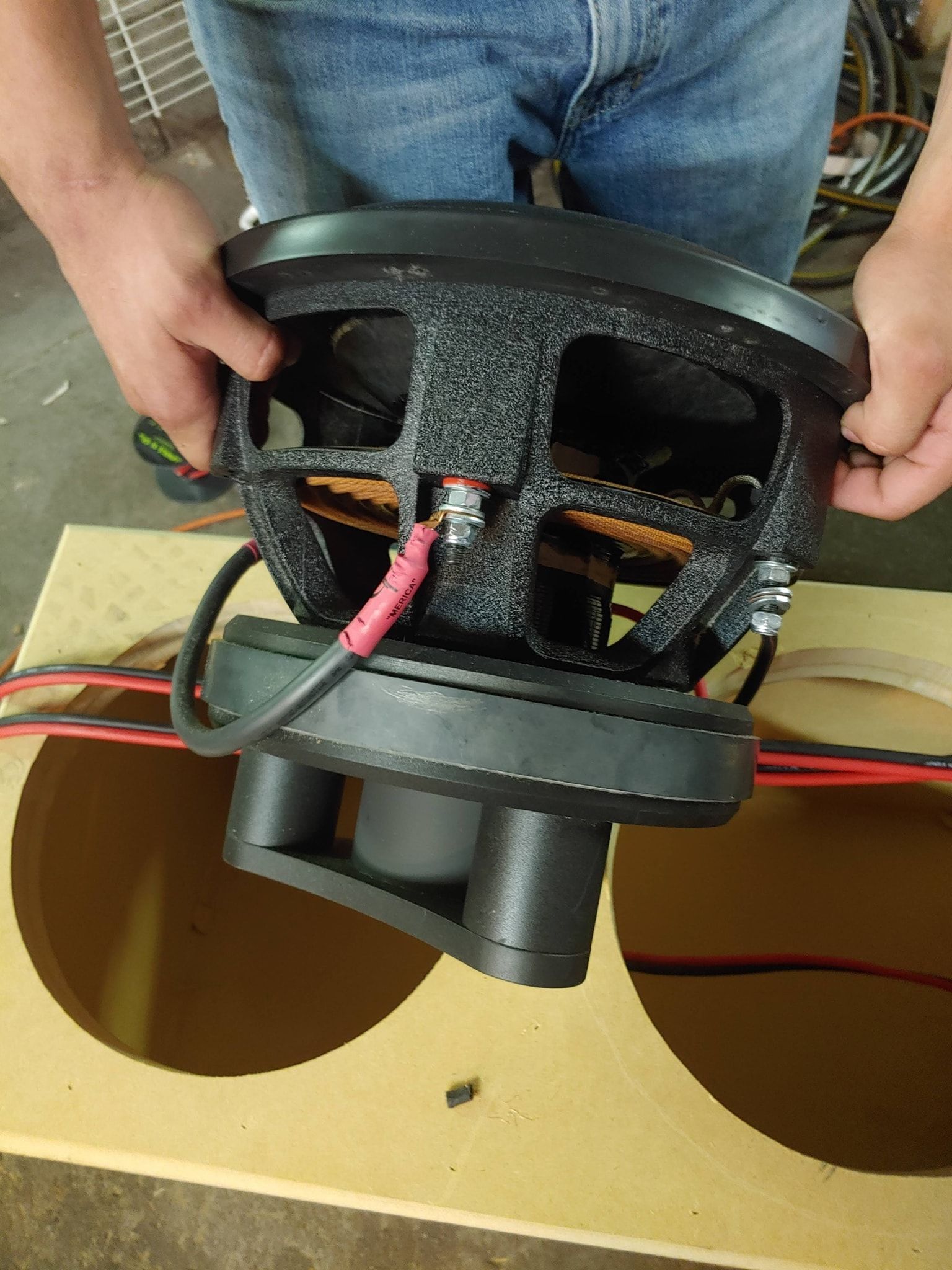 Person holding a black subwoofer speaker over a tan speaker box with red wires visible.