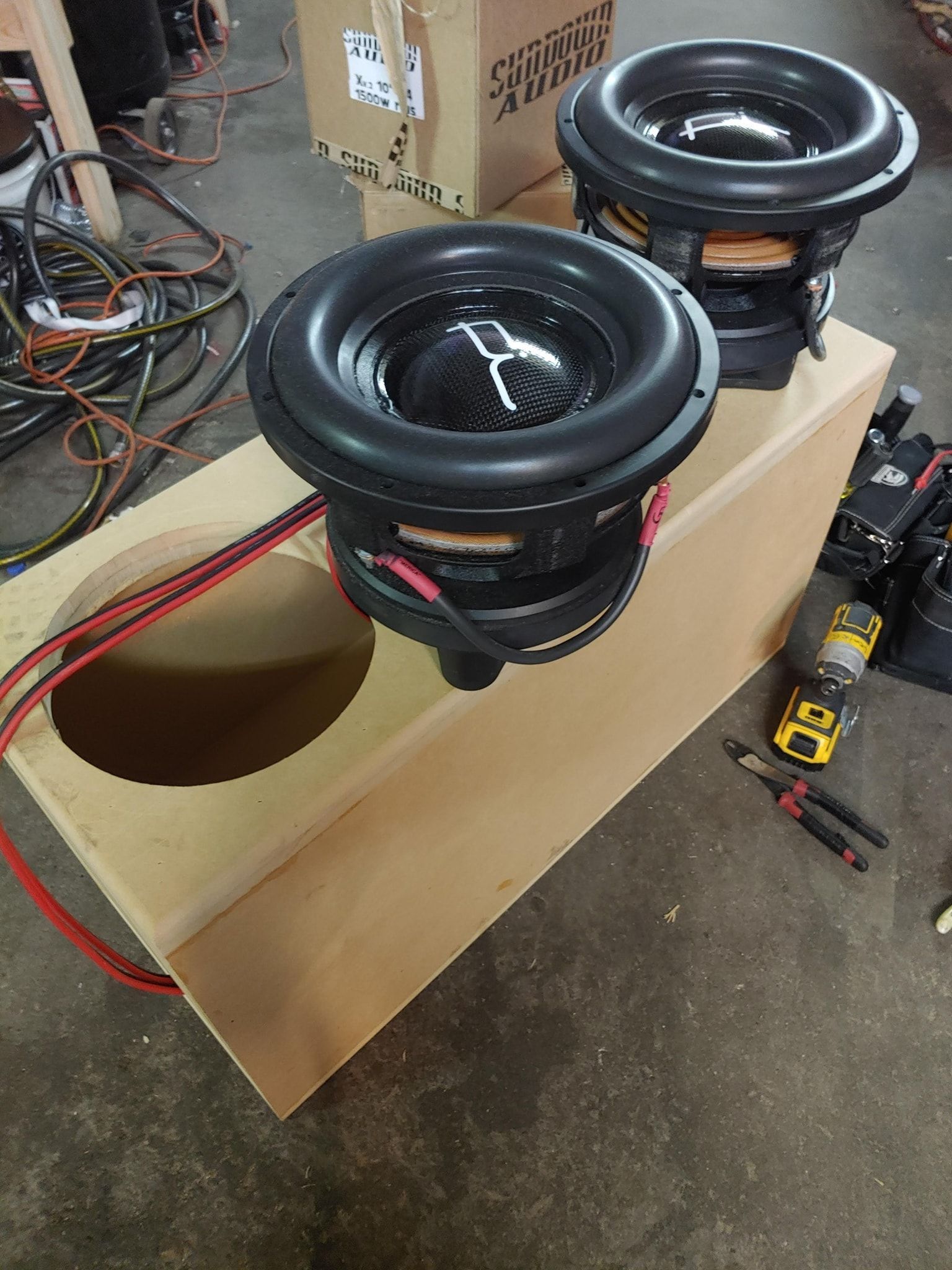 Two large subwoofers are mounted on a wooden enclosure in a workshop setting. Red wires are connected to the speakers.