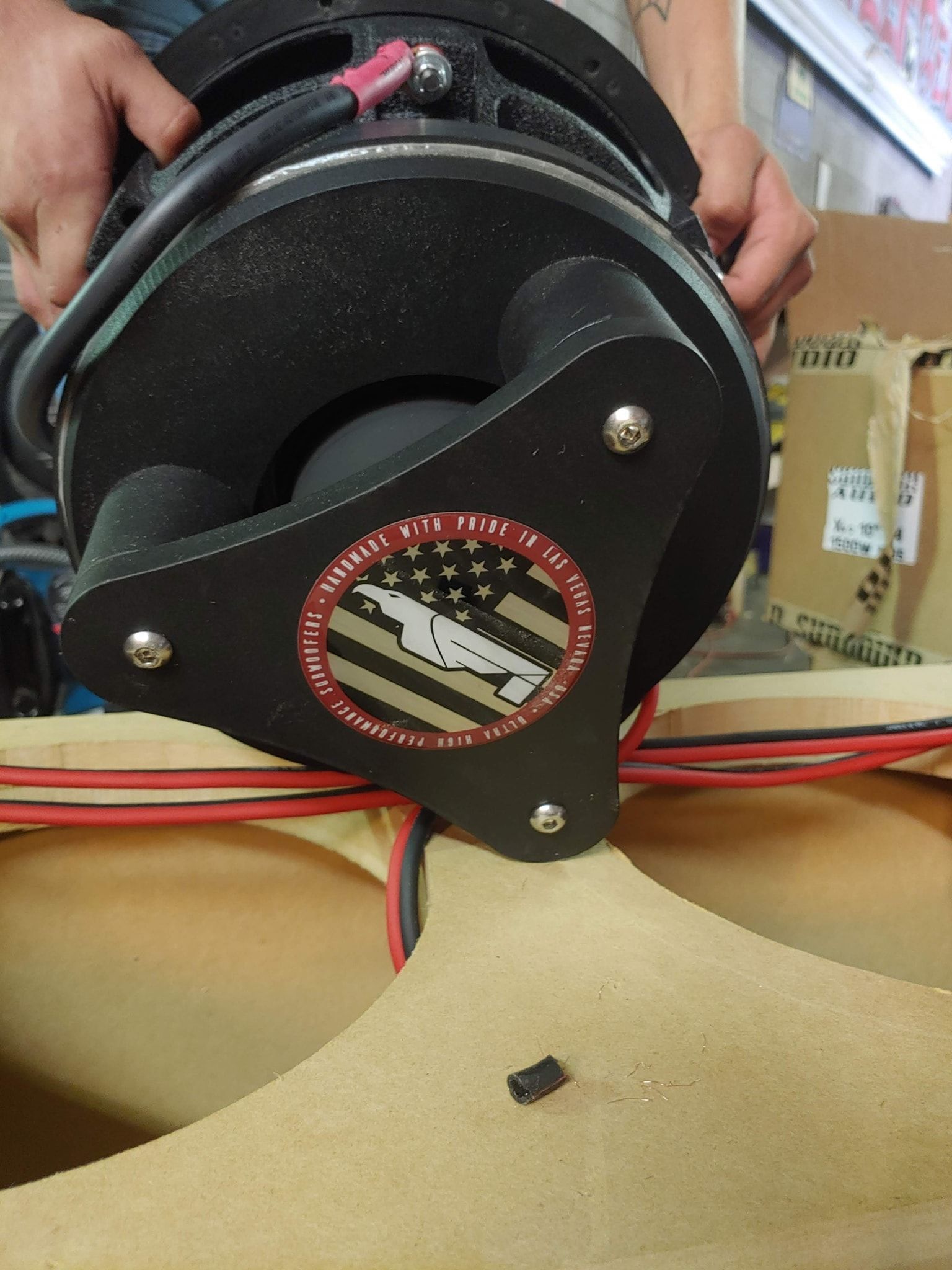 A person holds a large black subwoofer with a red, white, and blue emblem over a wooden speaker box.