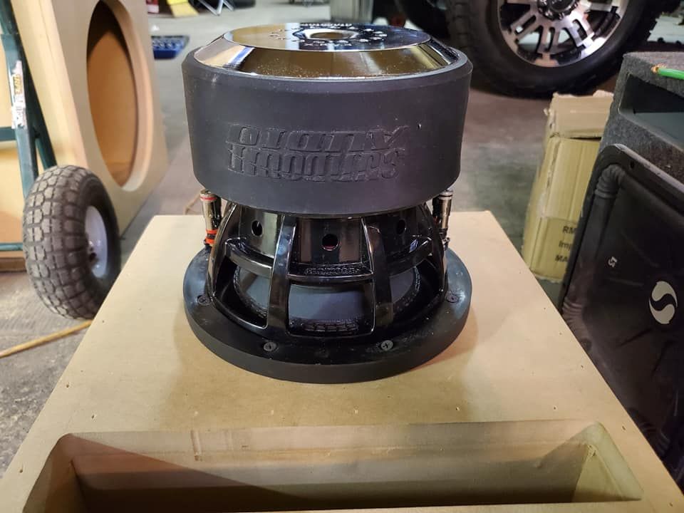 A Sundown Audio subwoofer sits on a beige box. Black metal frame, black foam surround, and a silver center are visible.