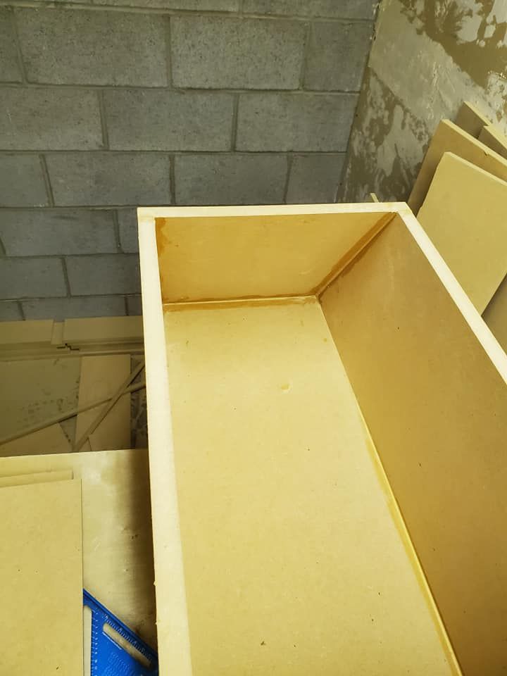 A rectangular wooden box, possibly a drawer, is partially assembled, with the interior visible against a cinder block wall.