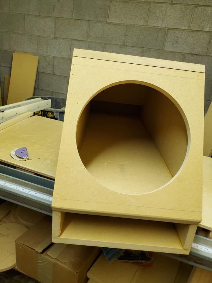 A wooden subwoofer box with a circular hole in the front, sitting on a workbench in a workshop.