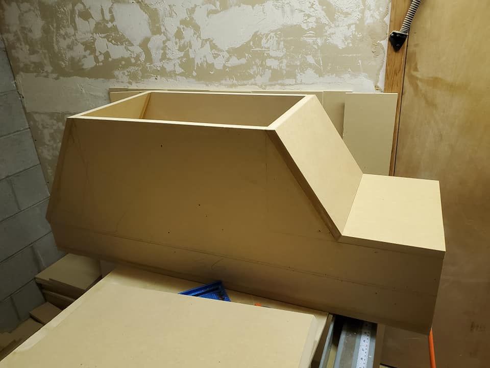 A beige wooden box with angled sides and a rectangular top, possibly a speaker enclosure, sitting on a workbench.