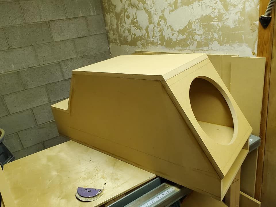 Assembled subwoofer enclosure made of tan MDF, with a large circular opening. It is on a workbench in a workshop.