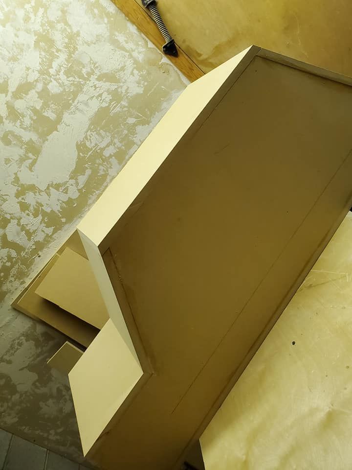 A partially constructed, painted cabinet in shades of cream and beige, against a rough wall.