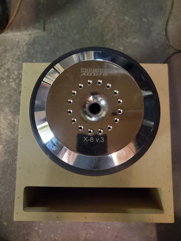 Sundown Audio X-8 v.3 subwoofer mounted in a tan wooden enclosure with a rectangular port.
