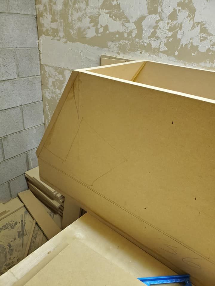 A wooden cabinet frame under construction, angled against a rough wall, showing drawn construction lines.