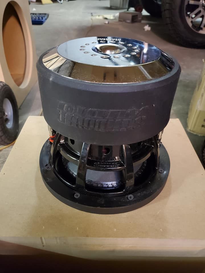A black and chrome subwoofer with a black rubber surround, sitting on a wooden surface in a workshop.