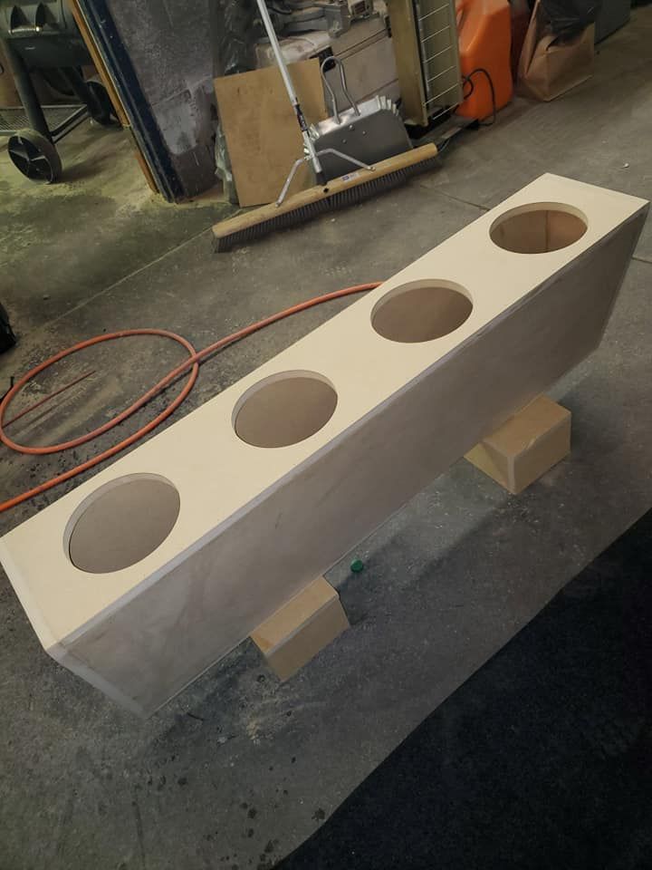 Unfinished wooden box with four large circular holes, likely for speakers. Resting on wood blocks in a workshop setting.