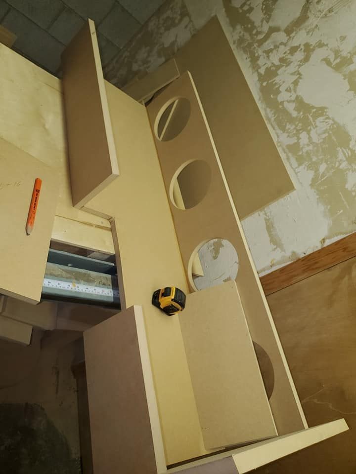 A DIY speaker box under construction, featuring plywood pieces with cut-out circles. A measuring tape rest on the wood.