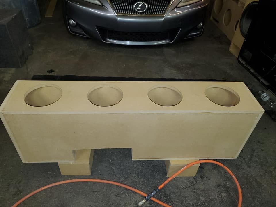 A beige subwoofer box with four speaker holes sits in front of a silver Lexus, likely being installed in a car audio setup.