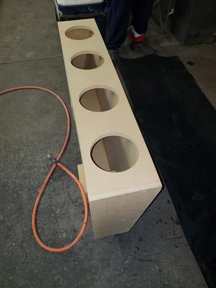 A rectangular wooden speaker box with four circular openings, positioned on a concrete floor.