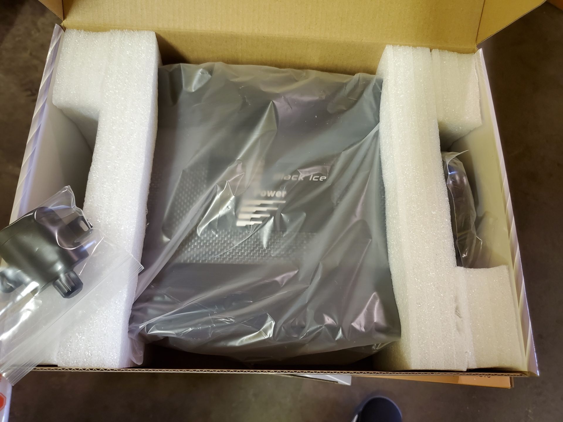 An open cardboard box containing a black electronic device wrapped in plastic, with foam padding. 