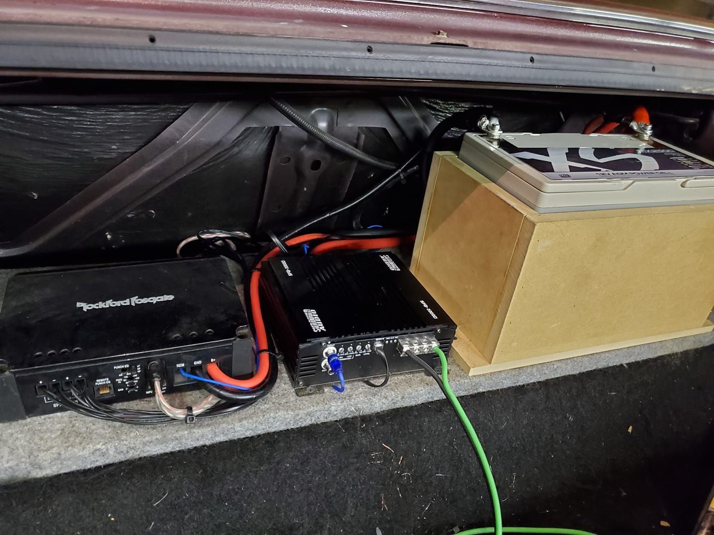 A car trunk with two audio amplifiers and a battery on a wooden box. Red and black wires connect the components.