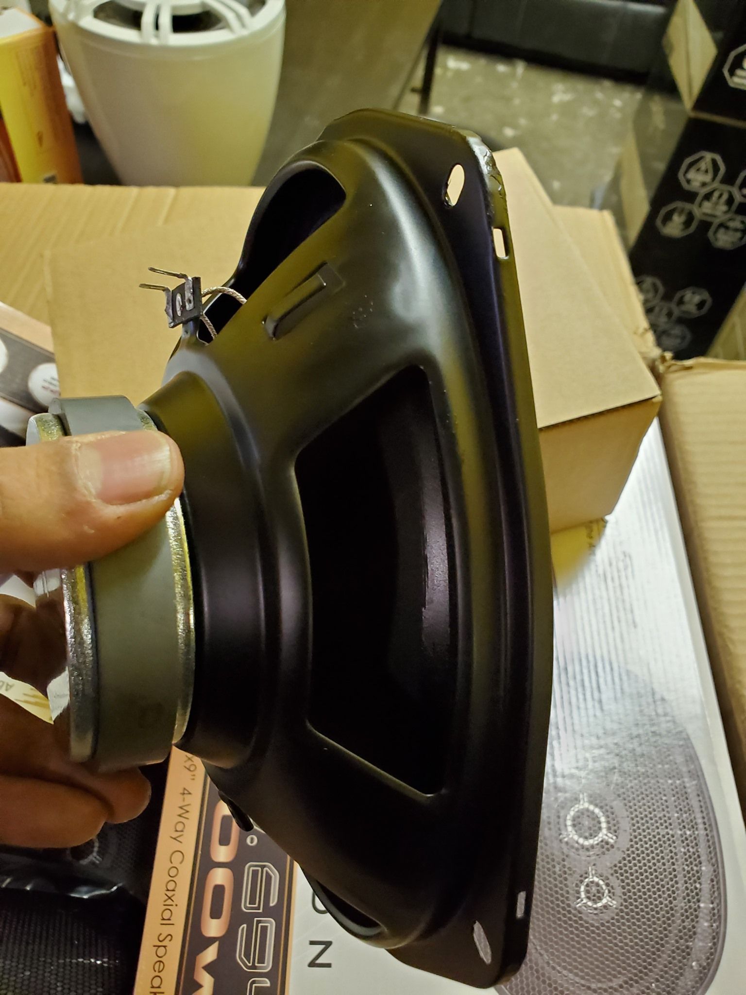 A person's hand holding a black speaker with a gray magnet, resting on cardboard.