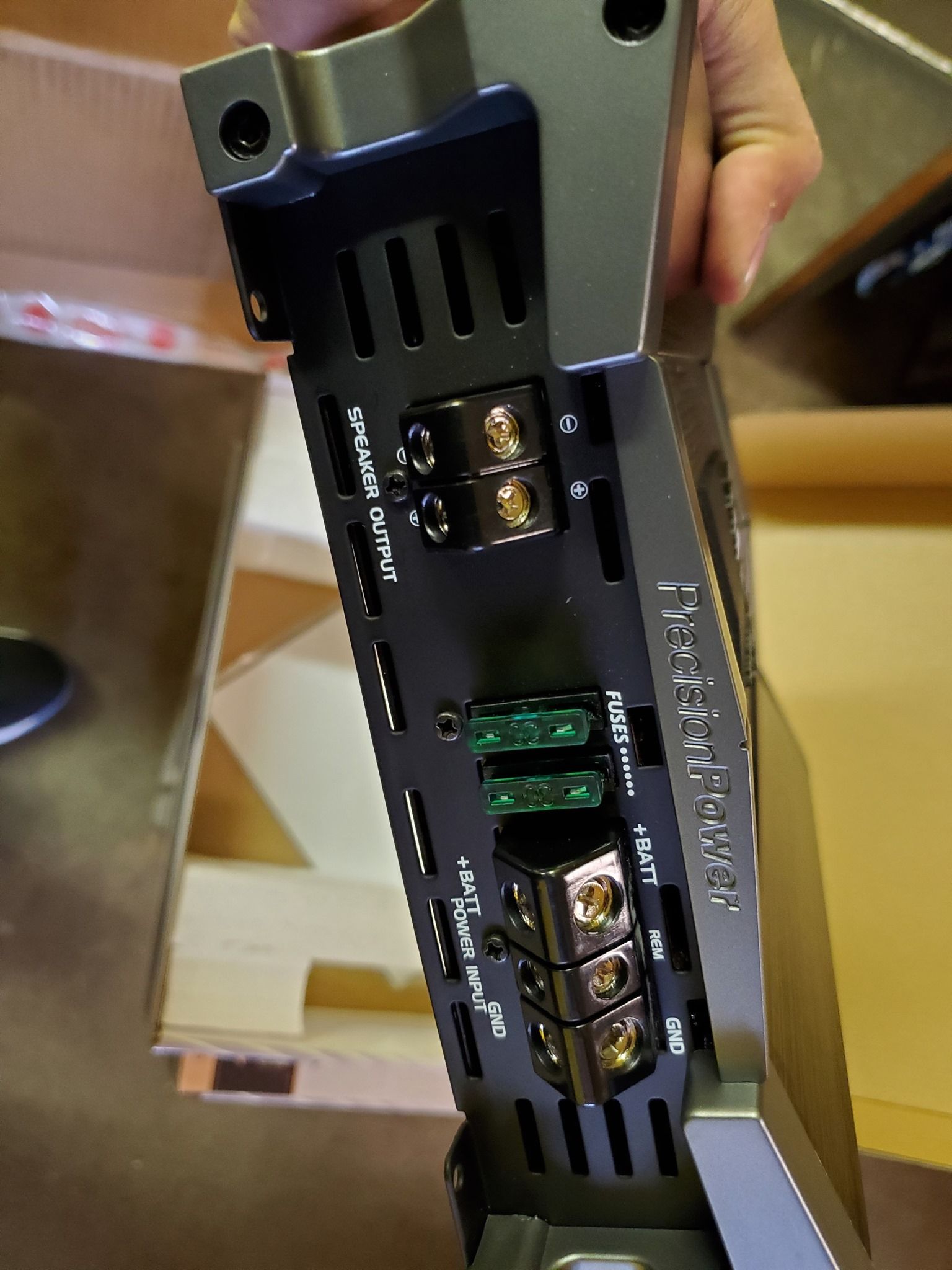 Close-up of a black car amplifier showing power and speaker terminals, along with two green fuses. 