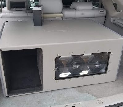 A custom car audio subwoofer enclosure in a car trunk. The enclosure is light gray with three visible subwoofers and a port.
