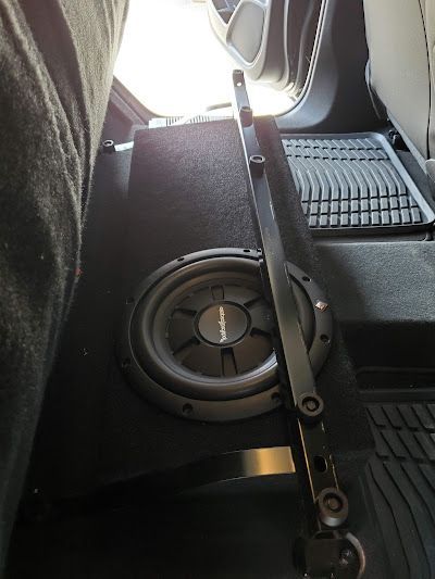 A car's interior showing a black subwoofer in a box behind the driver's seat. A black metal bracket is attached.