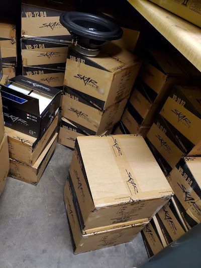 Boxes of car subwoofers stacked in a storage area; one speaker is placed on top. Brown cardboard, black speaker cones.