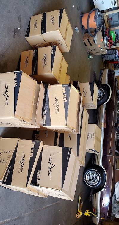 Boxes stacked on and around a classic car, likely audio equipment. Brown cardboard boxes with black and white labels.