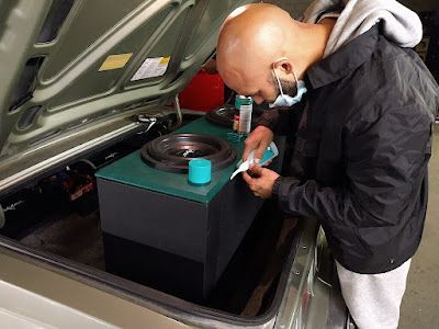 A man installs a subwoofer in a car trunk. He is wearing a mask and working on a green and black box.