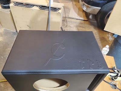 A black subwoofer box with an embossed logo on top, sitting in a workshop.