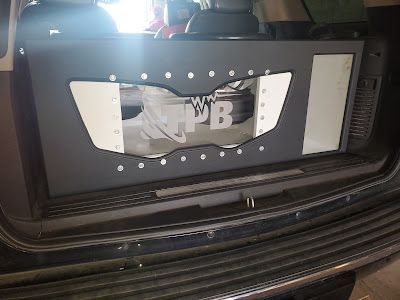 A custom subwoofer enclosure in the back of a dark SUV. The box is dark gray with a clear window and white accents.