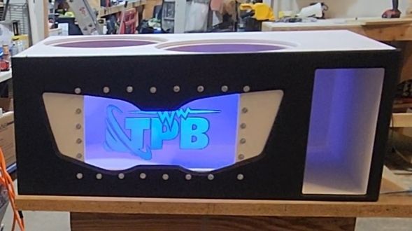 Custom car audio subwoofer box with two speaker holes and a lighted logo, plus a side window, on a workbench.