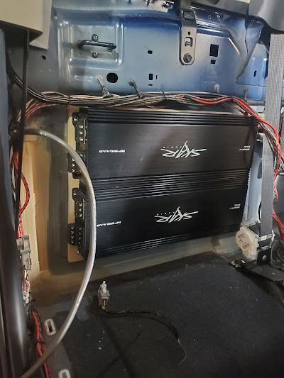 Two black car audio amplifiers stacked inside a vehicle, with wires running above them.