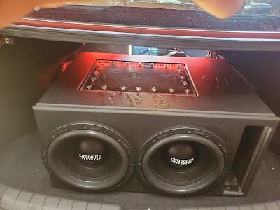 Two black subwoofers in a custom box with red lighting in a car trunk.